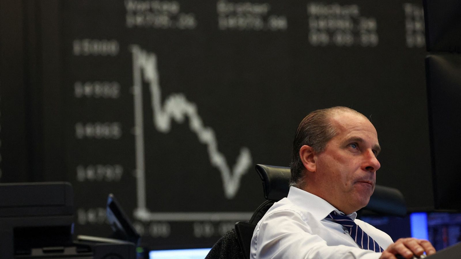 Un hombre trabaja en la Bolsa de Fráncfort frente a un ordenador y, de fondo, un gráfico refleja las caídas de los valores del DAX