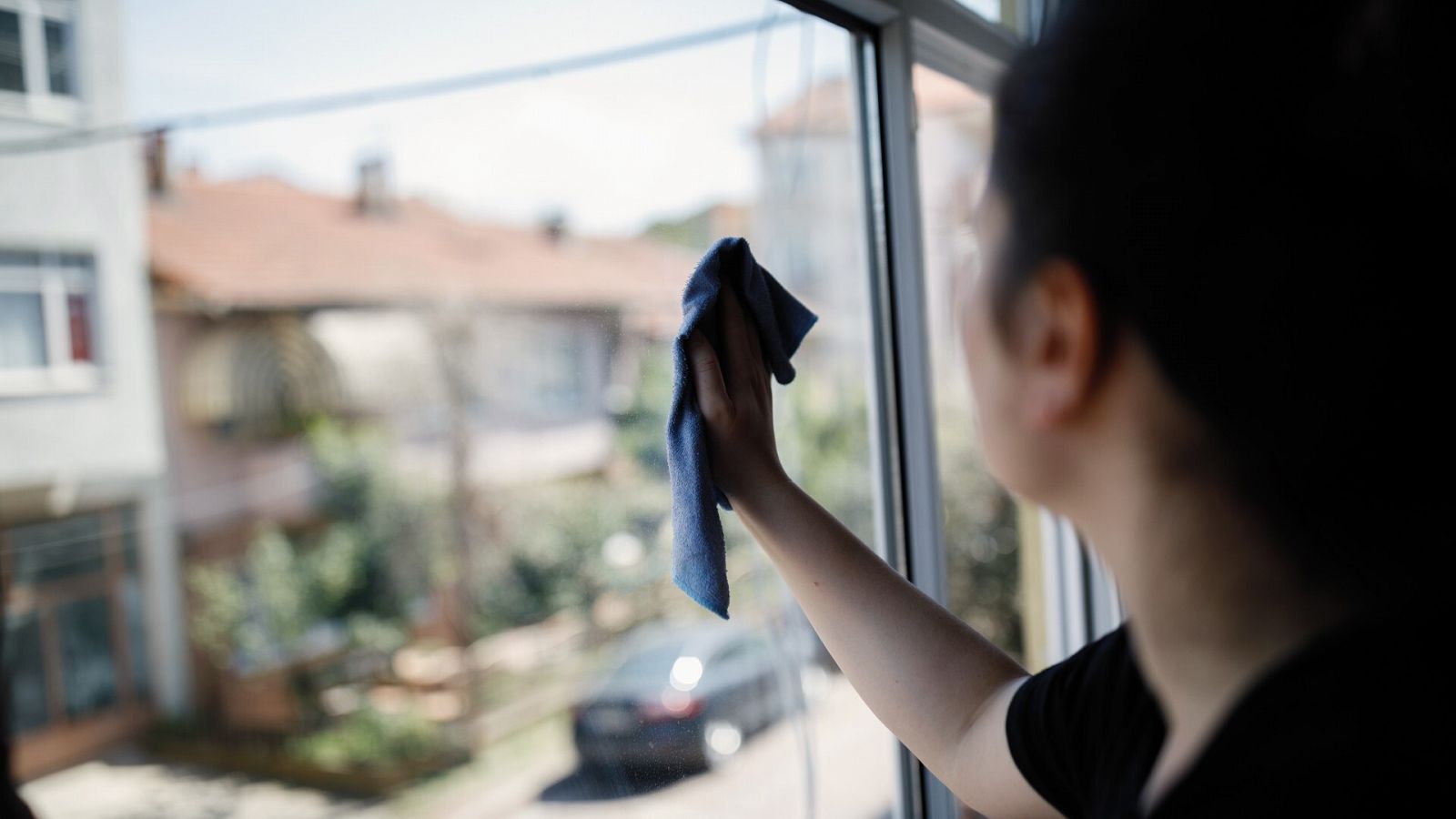 Una mujer joven limpia desde dentro el cristal de una ventana, en la que se ve la calle de un barrio periférico residencial.