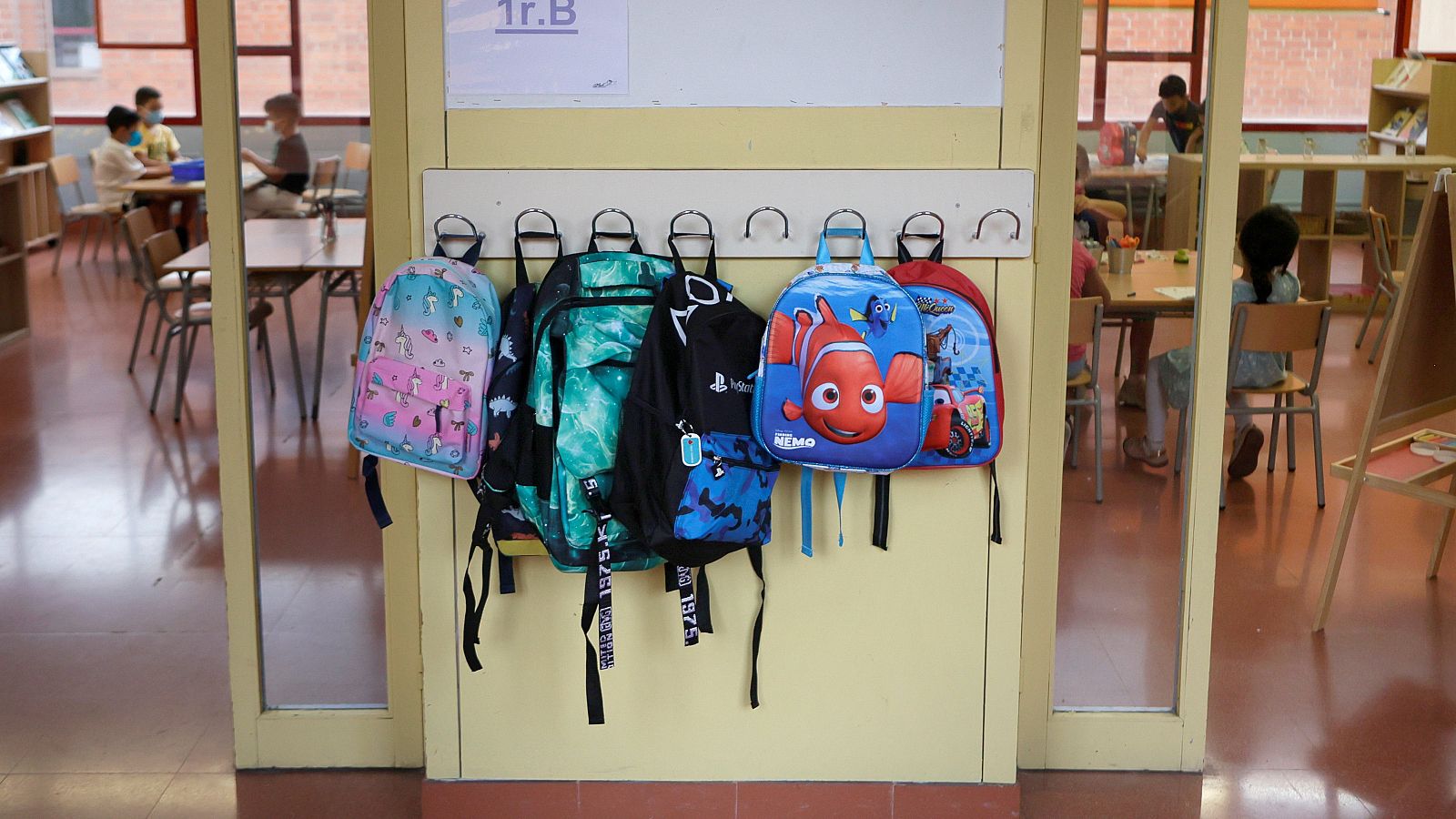 Entrada a un aula d eun colegio d eprimaria. En la pared se ven cuatro mochilas on dibujos animados decorativos colgadas de la pared.