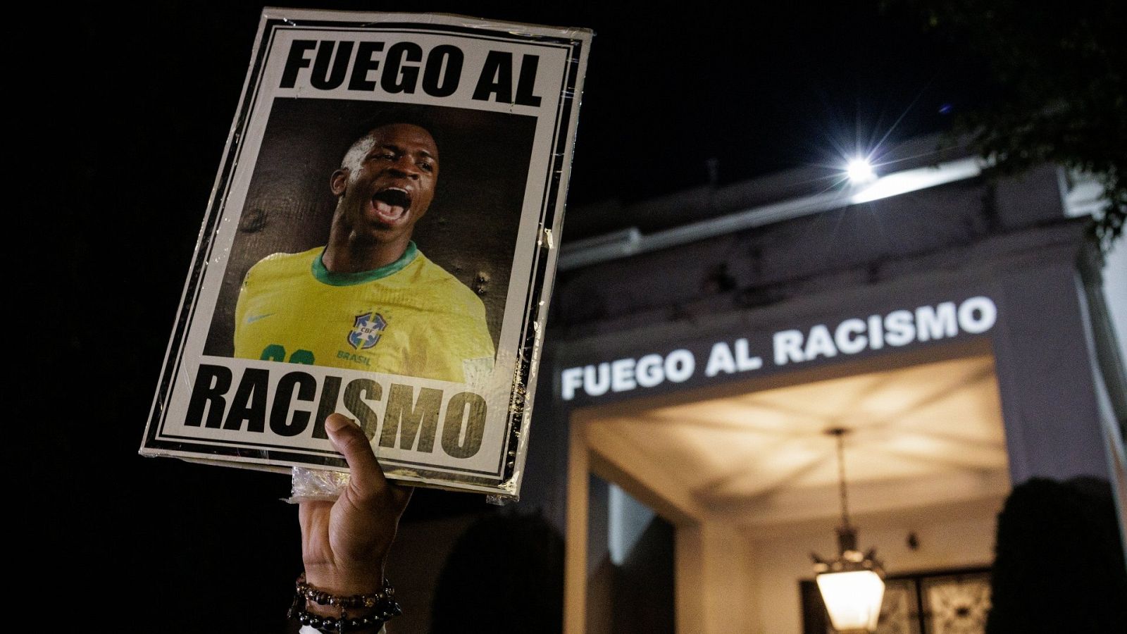 Una mujer sujeta en alto un cartel con la foto de Vinicius y un texto que dice "Fuego al racismo"