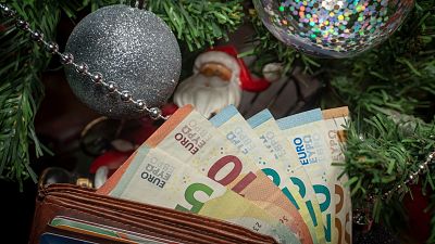 Árbol de Navidad decorado con bolas y un Papá Noel y en frente, unas manos sujetan una cartera llena de billetes