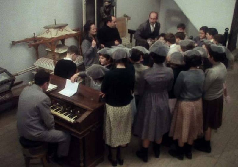 Lola y el parróco del pueblo enseñan a los niños una de las canciones que tienen que cantar en las fiestas del pueblo