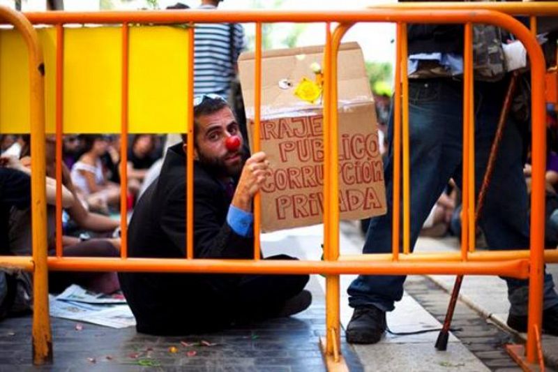 Un manifestante con naríz de payaso con una pancarta con el mensaje "traje público corrución privada".