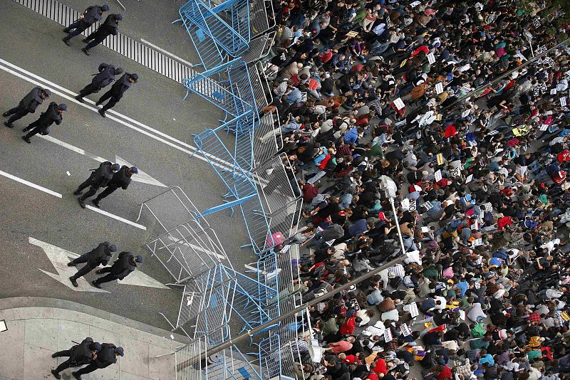 Protesters sit down as Spanish National Police officers in riot gear stand guard behind a fence closing the street outside Madrid's Parliament during a demonstration