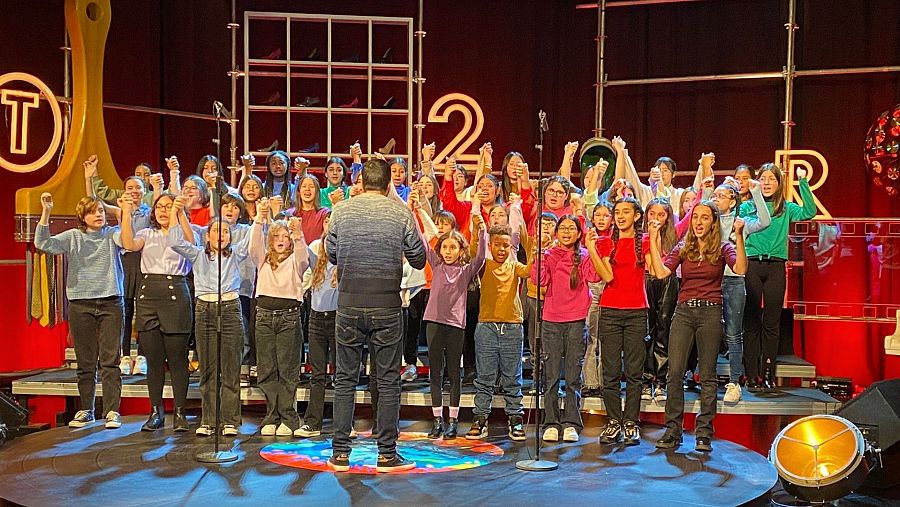 Coral de nenes i nens cantant a l'escenari del plató del programa amb el director d'esquenes