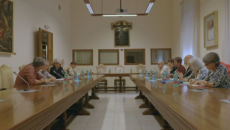 Reunión de hombres y mujeres en una sala presidida por un cuadro con un Cristo crucificado.