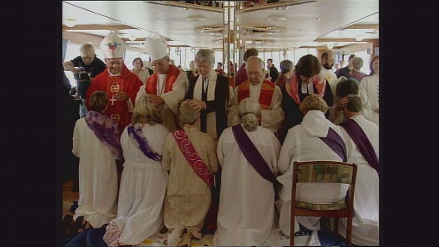 Obispos imponen las manos a mujeres arrodilladas mientras las ordenadan sacerdotes.