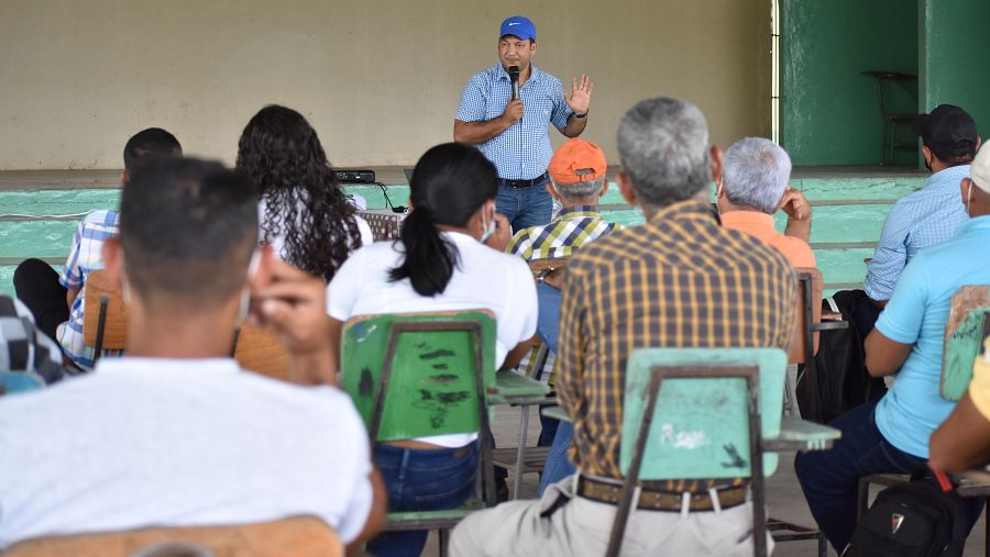 Hombre con gorra azul y micrófono se dirige a un auditorio de hombres y mujeres que aparecen de espaldas.