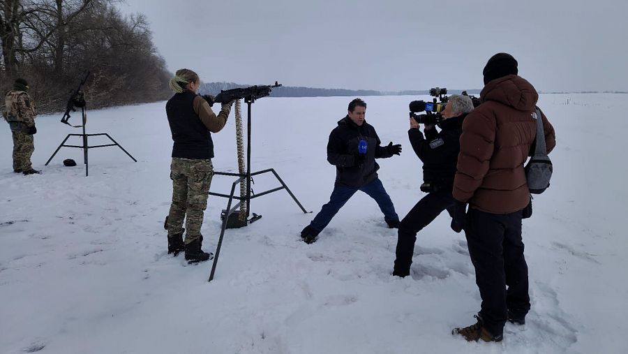 Un periodista con micrófono abre las piernas para estar más bajo y facilitar el encuadre del cámara junto al militar que está con una metralleta en un campo completamente nevado.
