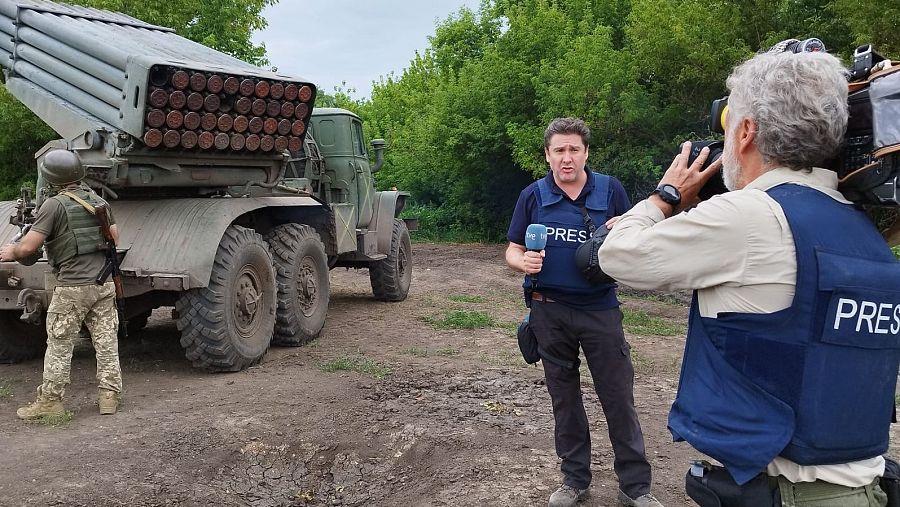 Un reportero con el micrófono de TVE, casco y chaleco, habla a cámara con un camión lanzamisiles a su espalda.