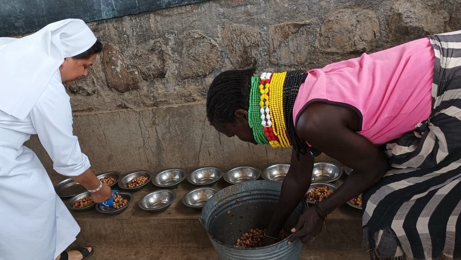 Una monja vestida de blanco y una mujer turkana con muchos collares de colores sirven comida en platos metálicos.