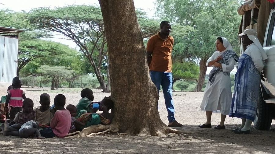 Un hombre conversa con dos misioneras junto al coche de estas mientras un grupo de niños están sentados bajo la sombra de un árbol.