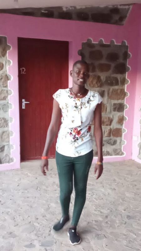 Una mujer africana de pelo corto con camisa blanca floreada y pantalón verde delante de una puerta rosa y una pared de piedra.