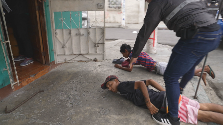 Dos jóvenes con indumentaria deportiva tumbados boja abajo en el suelo mienras un policía les cachea.