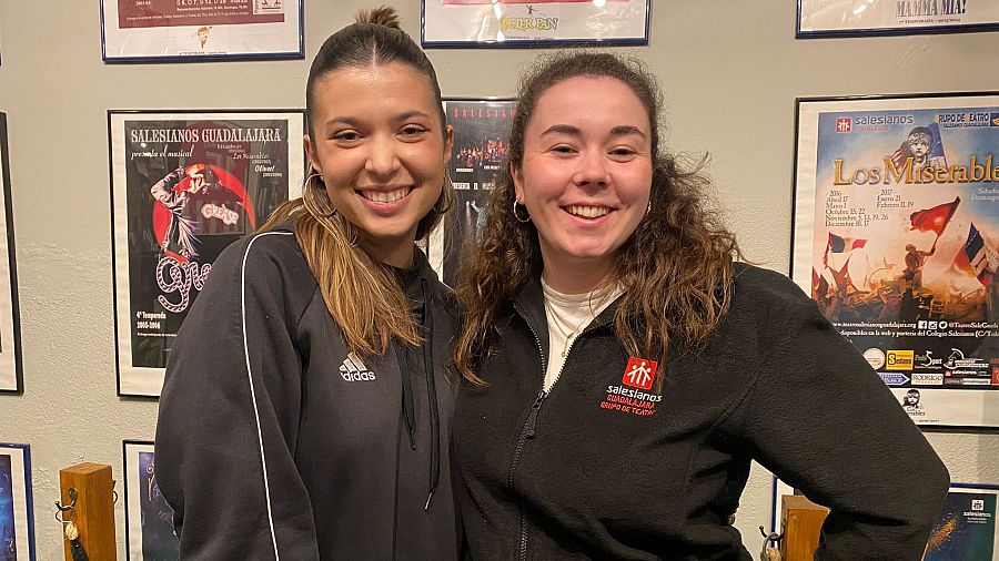 Dos chicas jóvenes sonríen a cámara con carteles enmarcados de musicales teatrales tras ellas.