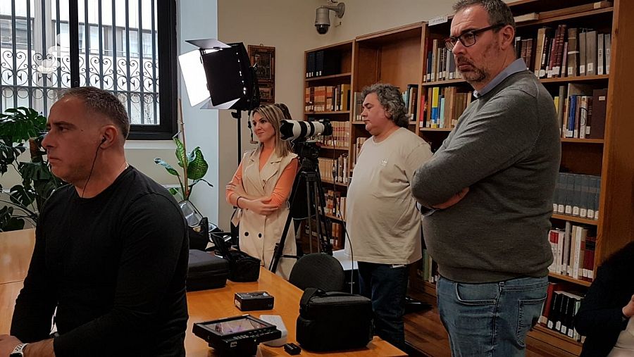 Personas grabando en una habitación con una estantería de libros detrás, una ventana, un foco y cámaras.