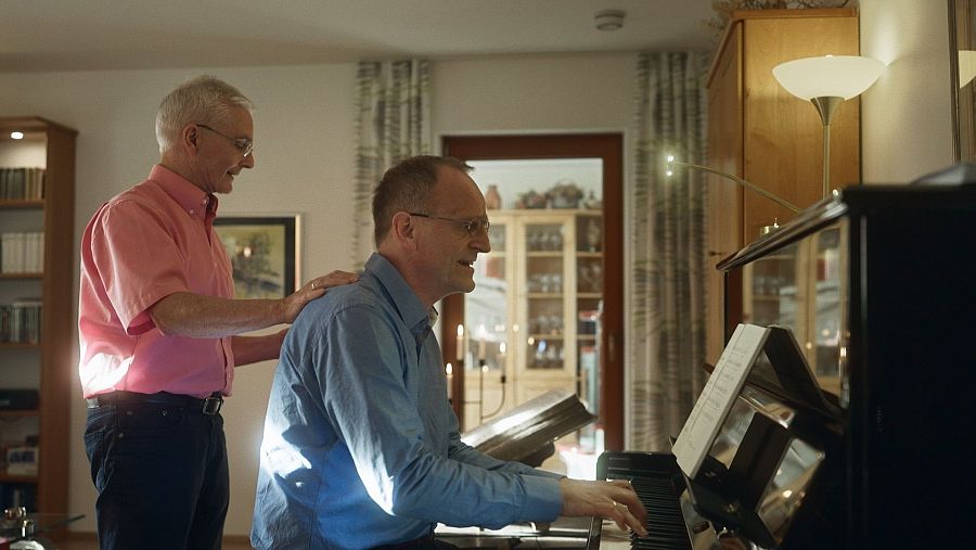 Un hombre toca el piano y otro apoya la mano derecha sobre la espalda del pianista