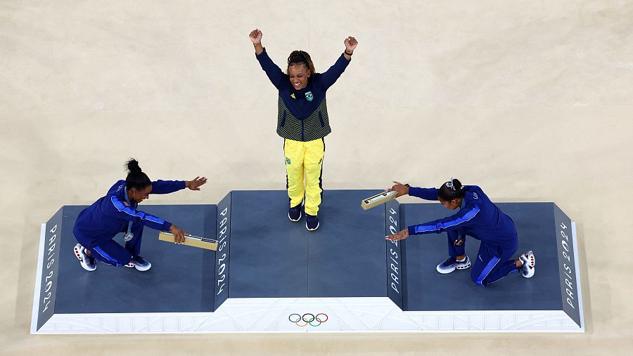 Rebeca Andrade celebra el oro olímpico en suelo mientras las estadounidenses Simone Biles y Jordan Chiles le hacen una reverencia.