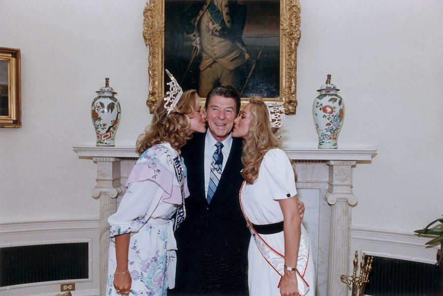 El presidente Ronald Reagan sonríe mientras Miss America, Kim Brede y Miss Universo, Shawn Weatherly, le dan un beso en ambas mejillas.