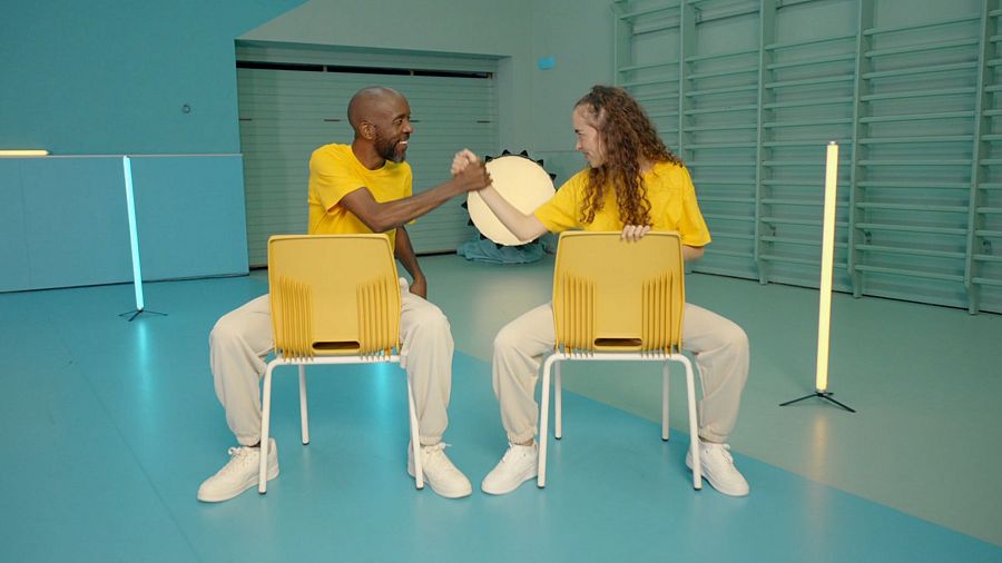 Martina Burón y Welelo Zamora se saludan dándose la mano, sentados en sillas amarillas en un estudio con una espaldera.