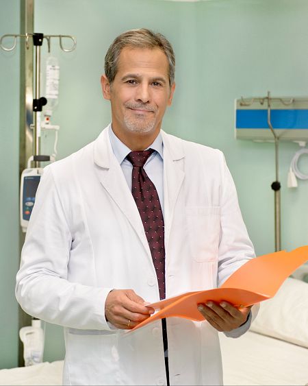 Médico con bata blanca y corbata granate revisa el historial de un paciente en una carpeta naranja en una habitación de hospital.