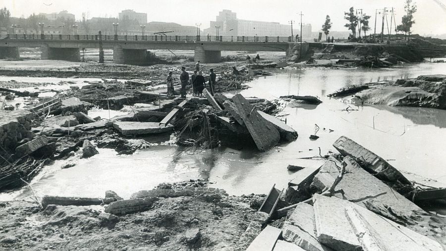 Escombros y personas inspeccionando los daños en el cauce del río Turia tras la Gran Riada de Valencia de 1957. Un puente se alza al fondo. No se identifican personajes públicos conocidos.