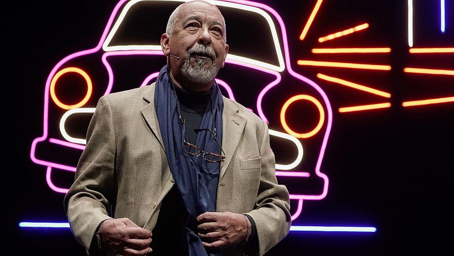 Leonardo Padura, reconocido autor cubano, posa en Oviedo durante un evento cultural, con un fondo de luces de neón que representan un coche.