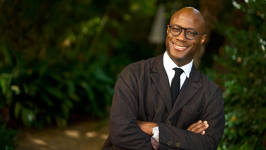 Barry Jenkins, con los brazos cruzados, sonríe a la cámara en un entorno ajardinado.
