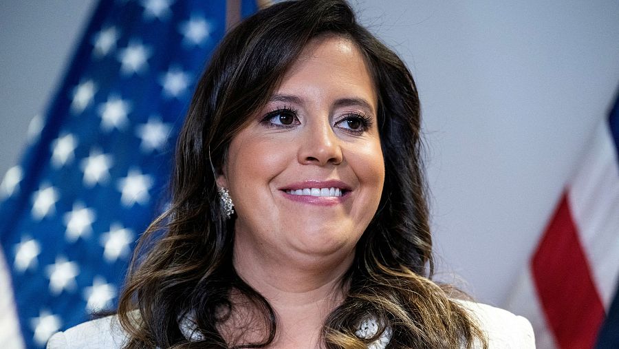 La congresista republicana estadounidense Elise Stefanik mira con una leve sonrisa a la derecha de la imagen, con la bandera de Estados Unidos desenfocada al fondo.