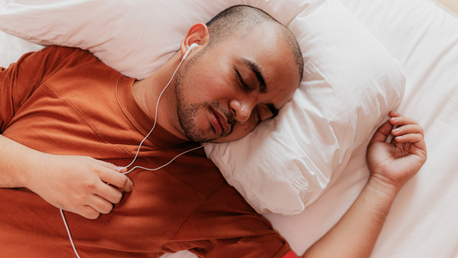 Hombre con camiseta naranja duerme con auriculares puestos en una cama.