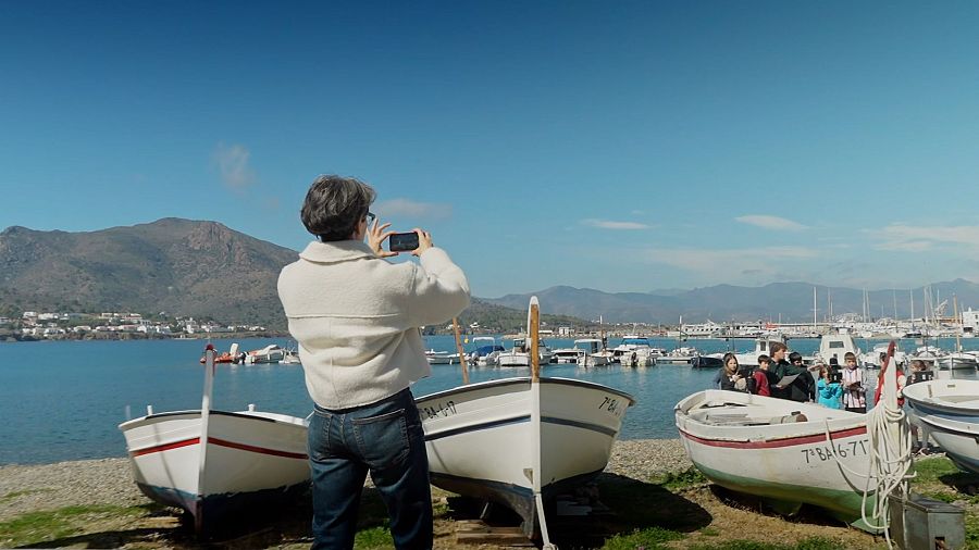 Mujer fotografiando un puerto con barcos y un grupo de niños con tablets, con montañas al fondo.