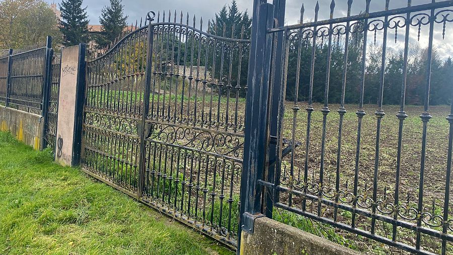 Verja metálica cerrada con un edificio al fondo y un terreno con vegetación al otro lado.