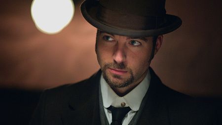 Víctor Ros, vestido de época, mira con seriedad hacia la derecha en un ambiente con luz tenue.