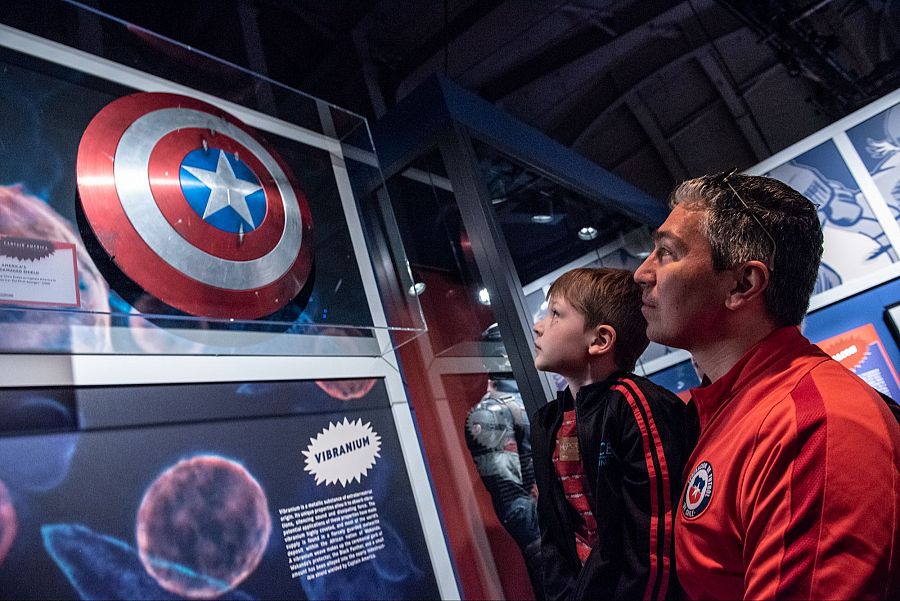 Un niño y un adulto observan el escudo del Capitán América en una exposición sobre el universo Marvel.