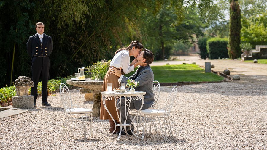 En los jardines de La Promesa, una pareja comparte un beso romántico mientras un mayordomo observa.