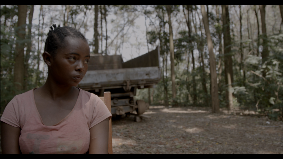 Joven sentada en primer plano, con un camión y un bosque al fondo.