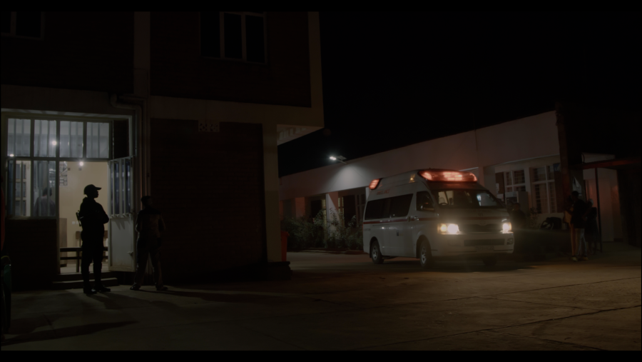 Una ambulancia llega a un edificio por la noche, con personas esperando en la entrada y cerca del vehículo.