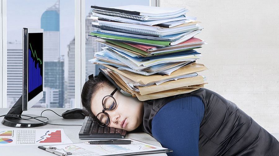 Mujer con gafas duerme en el teclado, agobiada por una pila de papeles, ilustrando el estrés laboral.