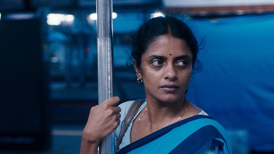 Mujer en un autobús agarrada a una barra metálica, mirando a la derecha. Lleva un sari azul.