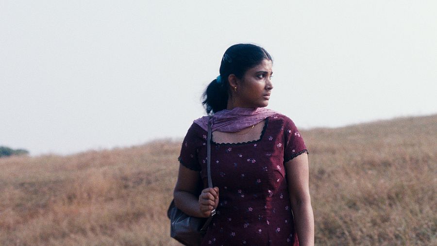 Una mujer con vestido burdeos y pañuelo malva camina por un campo con la mirada fija en el horizonte.