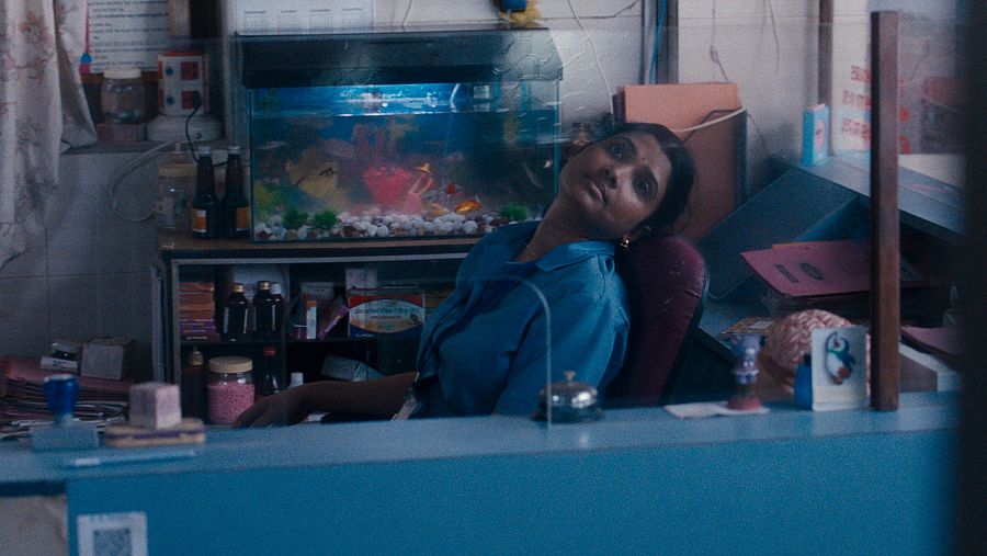 Una mujer con uniforme azul mira hacia arriba pensativa, tras el mostrador de una oficina con una pecera en la pared.