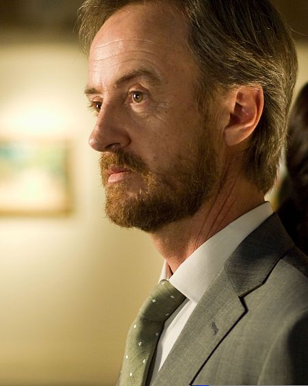 El actor Fernando Cayo, con traje gris y corbata verde, mira seriamente hacia la izquierda.