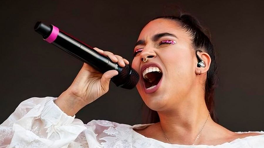 María José Llergo canta con intensidad en una actuación, con micrófono en mano y blusa blanca.