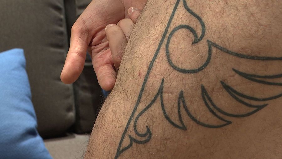 Primer plano de un tatuaje en una pierna, que parece una pluma o un ala, mientras una mano lo toca.
