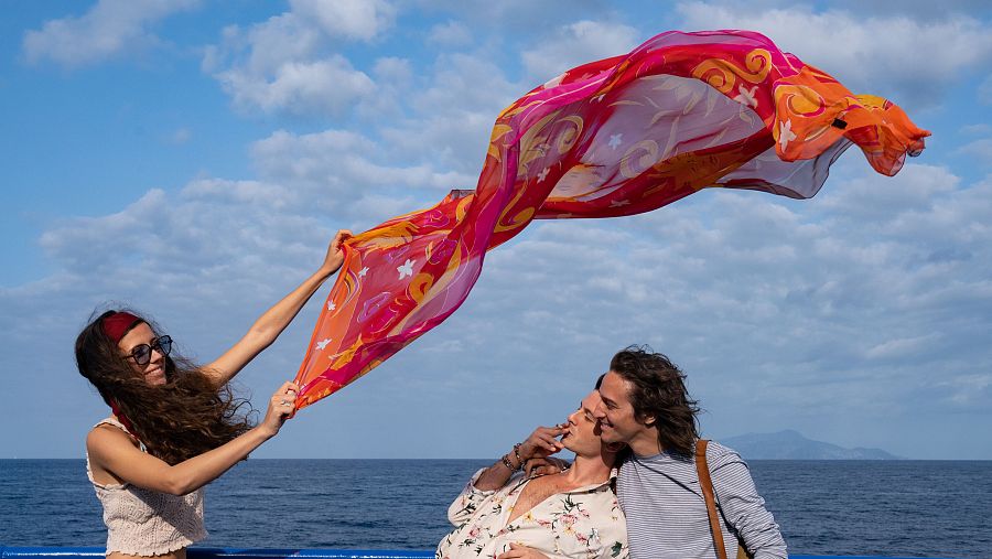 Pareja disfruta de un día soleado en la cubierta de un barco, con el mar y una isla al fondo.