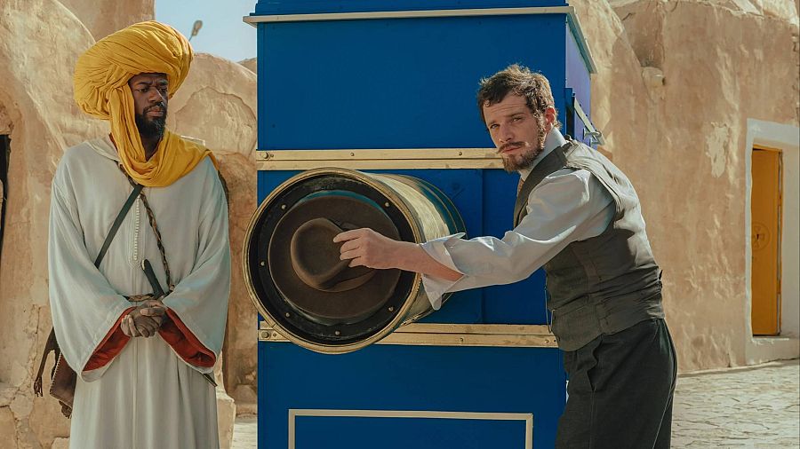 Un hombre introduce un sombrero en un aparato azul mientras otro con turbante amarillo lo observa en un ambiente desértico.