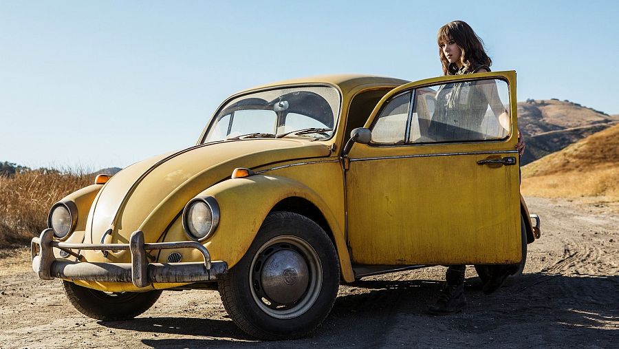 Hailee Steinfeld en una escena de *Bumblebee*, junto a un Volkswagen Beetle amarillo clásico en un camino de tierra.