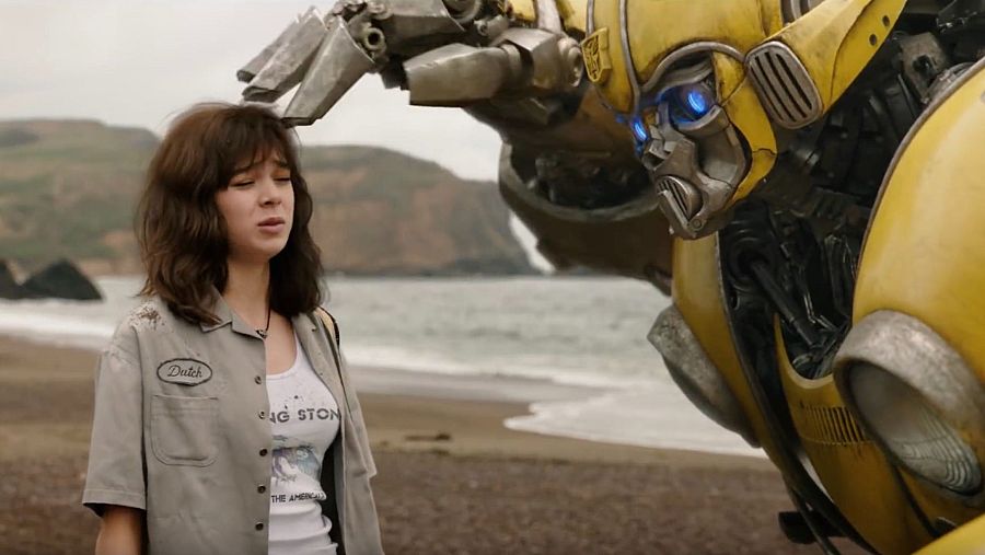 Hailee Steinfeld en una escena de playa de la película 'Bumblebee'.  Interactúa con el robot amarillo. Lleva camiseta blanca y camisa gris.