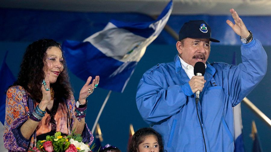 Ortega y Murillo en acto público. Ortega habla por micrófono; Murillo aplaude. Banderas de Nicaragua presentes.