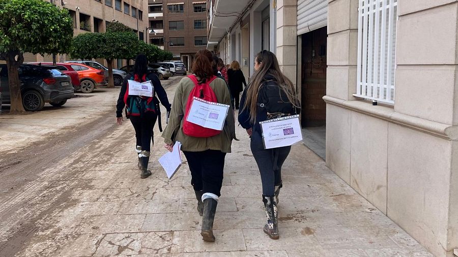 Mujeres de la Asociación Alanna realizan labor social puerta a puerta, llevando mochilas y carteles informativos. Trabajo de campo en una calle con coches aparcados.